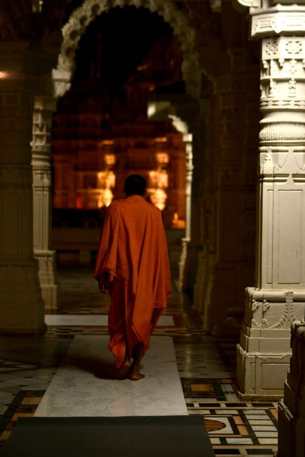 A person dressed in traditional saffron robes is walking along a polished white marble corridor inside the Neasden Temple, with intricately carved stone pillars on either side. The corridor is illumin