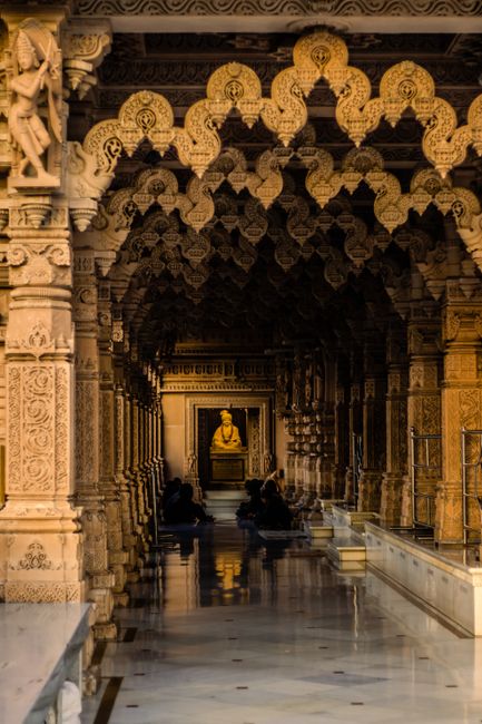 A person dressed in traditional saffron robes is walking along a polished white marble corridor inside the Neasden Temple, with intricately carved stone pillars on either side. The corridor is illumin